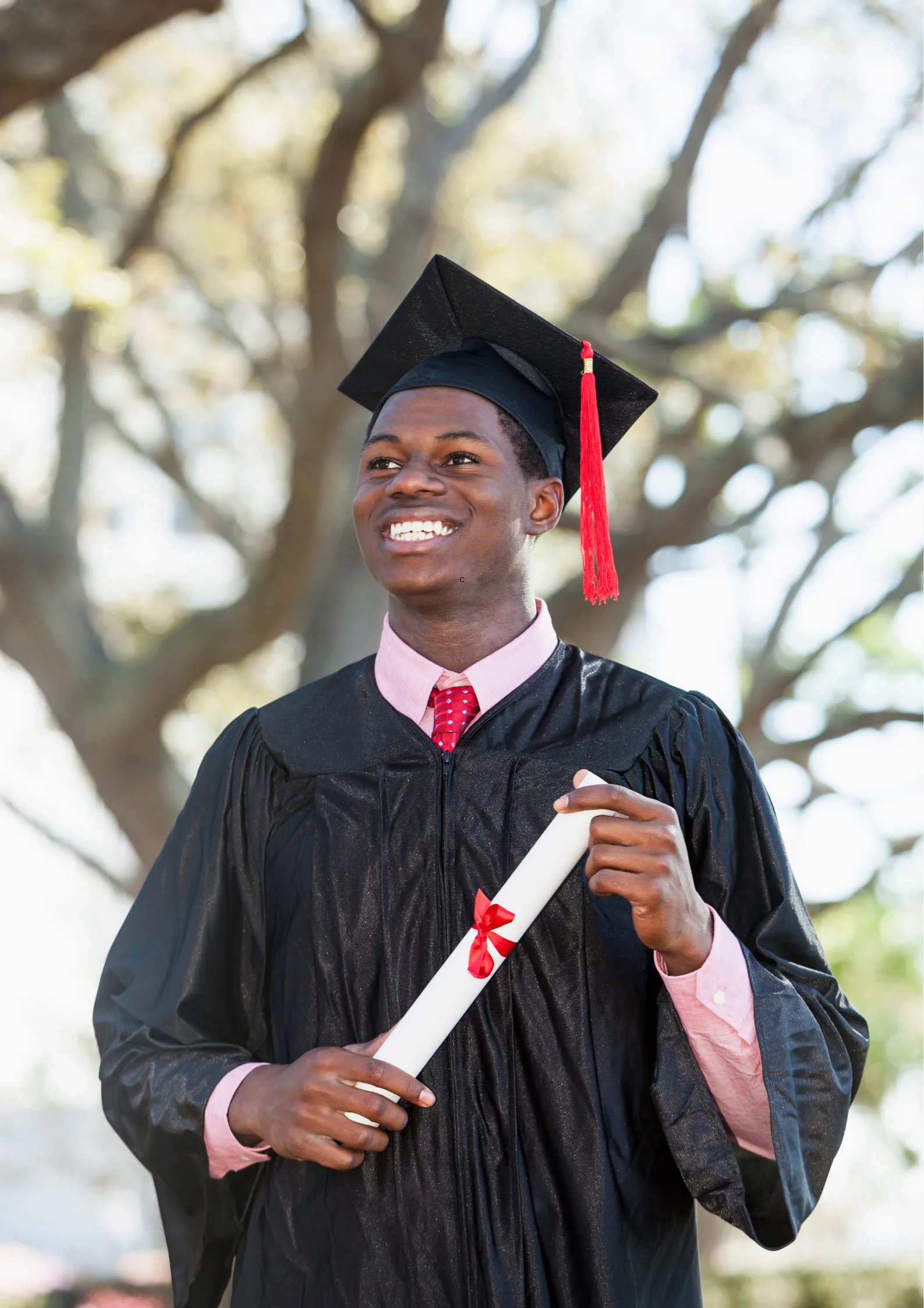 A smiling young man wearing a graduation cap and gown, holding a diploma tied with a red ribbon, standing outdoors on a sunny day.